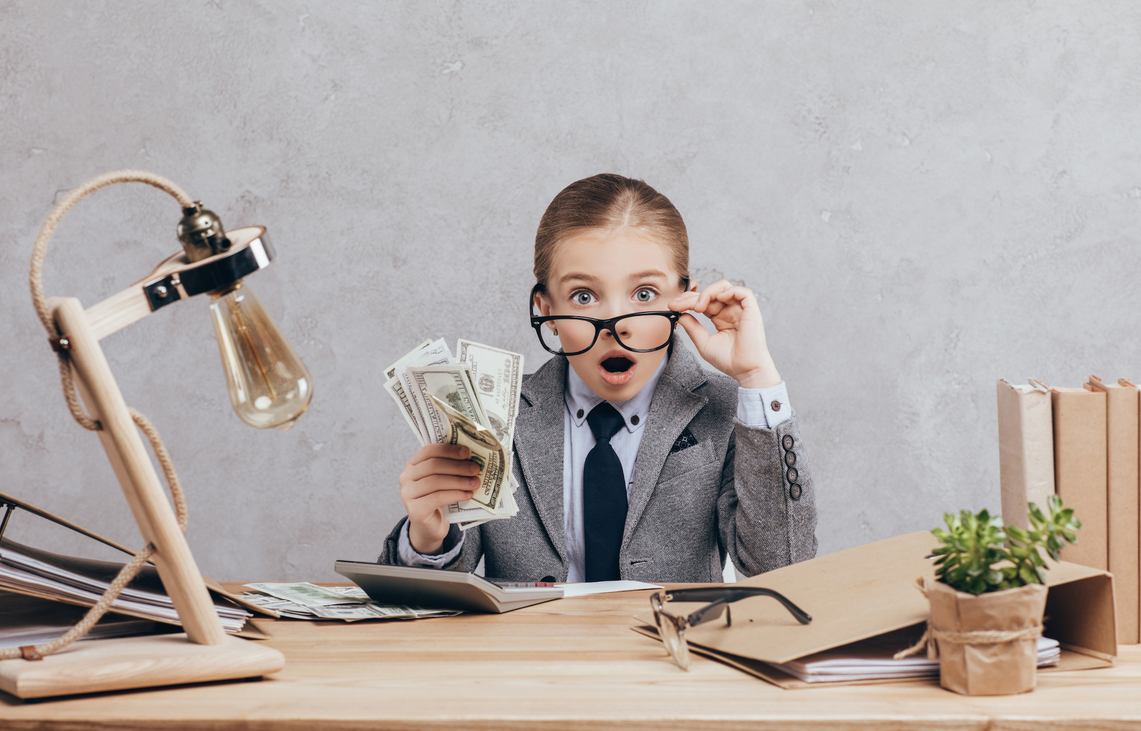 Young child in a suit holding dollar bills at a desk, representing early money lessons for kids.