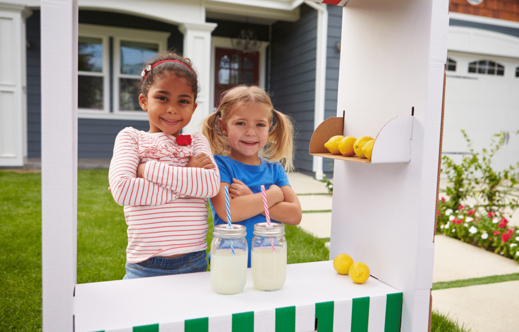 Two young girls stand proudly at their lemonade stand, showing how kids can learn entrepreneurship through hands-on business activities.