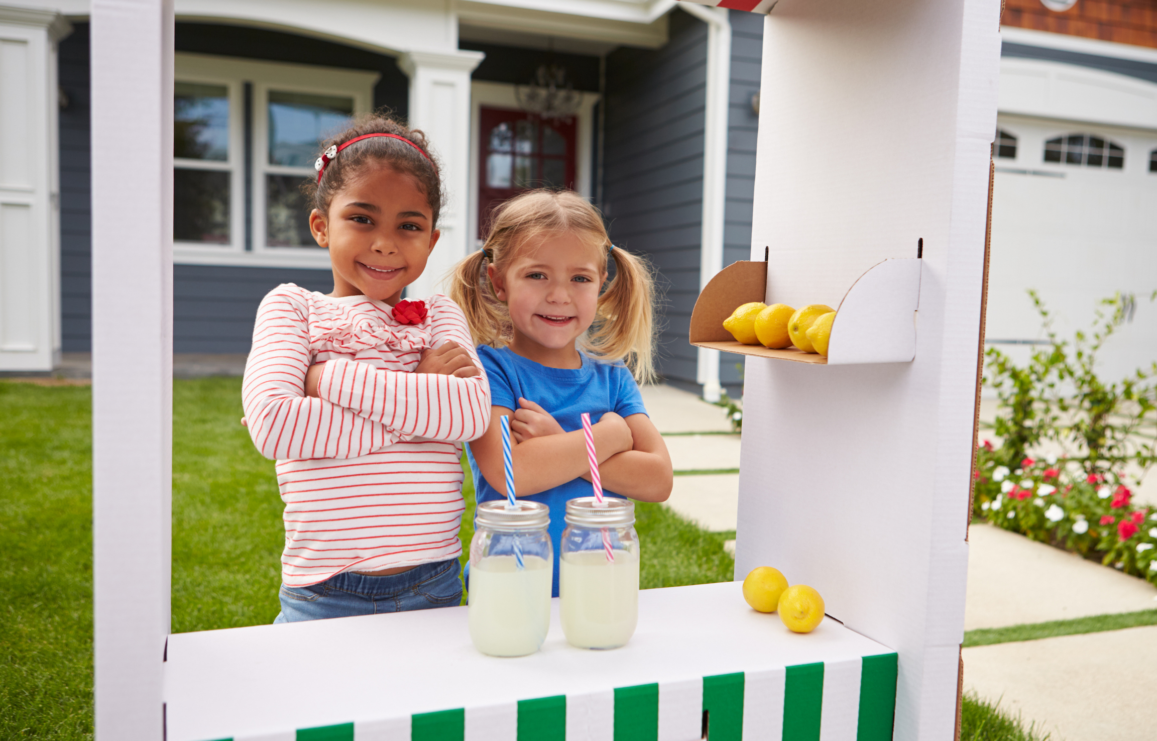 Two young girls stand proudly at their lemonade stand, showing how kids can learn entrepreneurship through hands-on business activities.